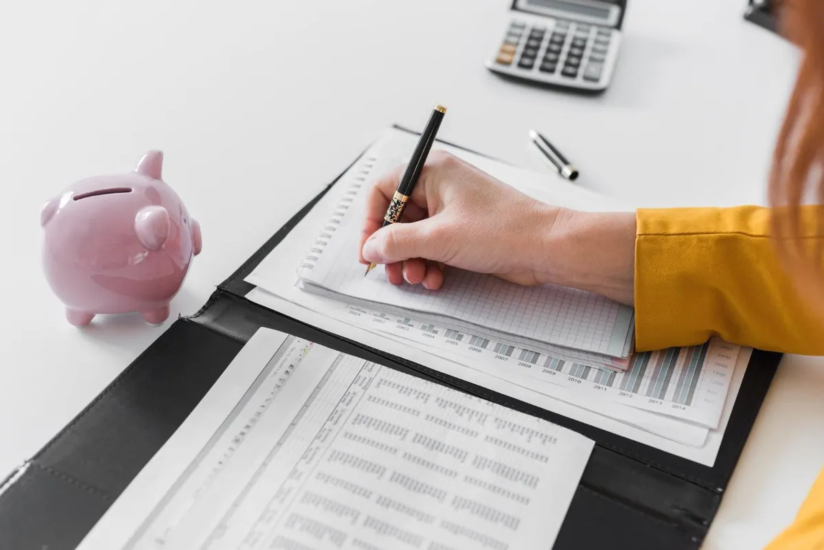 Persona anotando en una carpeta con planillas y gráficos, junto a una calculadora y una alcancía