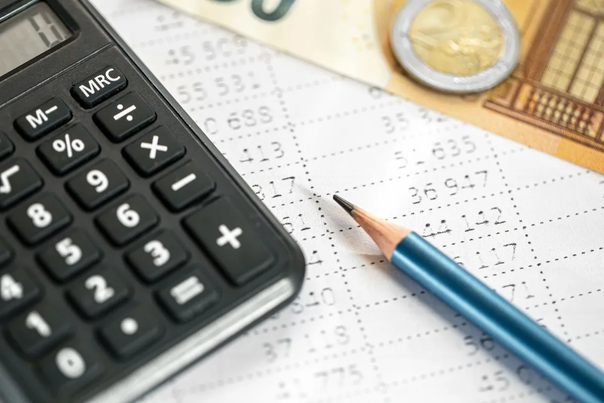 Calculadora y lápiz sobre una planilla de números, con billetes y monedas de fondo